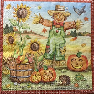 May include: A decorative napkin featuring a fall harvest scene. The image includes a smiling scarecrow in overalls, sunflowers, pumpkins with carved faces, and a wooden bucket filled with apples. The border is a stitched red design.