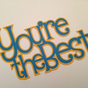 May include: A paper cutout with the phrase "You're the Best" in a cursive font. The text is blue with a yellow outline, set against a white background. Suitable for crafting or as a gift.