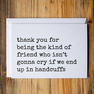 May include: A white greeting card with the text "thank you for being the kind of friend who isn't gonna cry if we end up in handcuffs" in black font. The card is inside a white envelope, set against a wooden background.