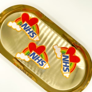 May include: Embroidered patches with a rainbow, red heart, and the letters "NHS" in blue. The patches are displayed on a gold-coloured oval tray. The rainbow has multiple colours, and the heart is a solid red colour.