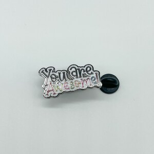 May include: A silver enamel pin with the text "You are awesome!" in rainbow colors. The pin has a black rubber backing.