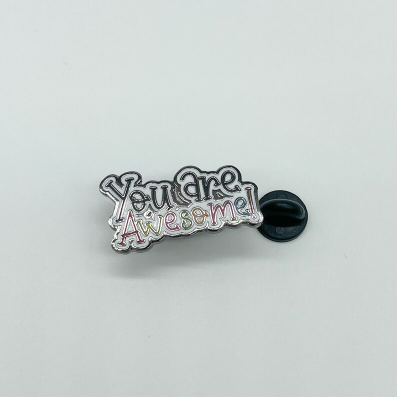 May include: A silver enamel pin with the text "You are awesome!" in rainbow colors. The pin has a black rubber backing.