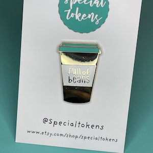 May include: Gold-coloured enamel pin shaped like a coffee cup with a teal lid. The cup has a white band with the text "full of beans" on it. The pin is on a white card with the text "@SpecialTokens" and "www.etsy.com/shop/specialtokens".