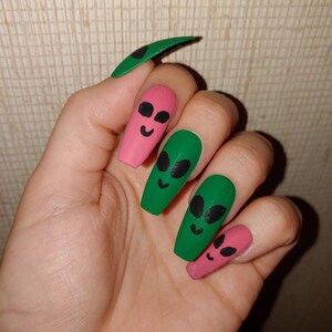 May include: Close-up of a hand with five long, fake nails. Two nails are painted green with black alien faces, and three nails are painted pink with black alien faces.