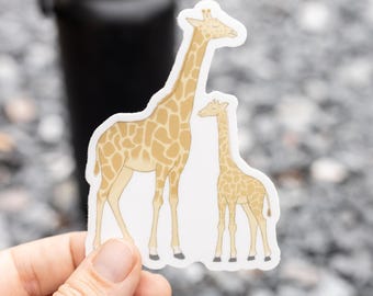May include: A sticker featuring two giraffes, one adult and one juvenile. The giraffes have tan fur with brown spots and are set against a white background. The sticker is held in a hand, with a blurred background.
