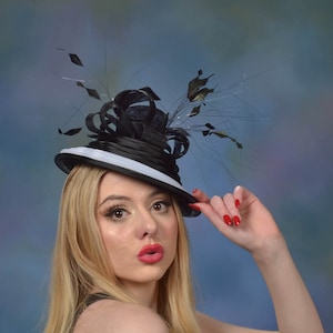 Puede incluir: Una mujer con un sombrero blanco y negro con una pluma negra y un vestido negro. El sombrero tiene un ala ancha y una banda blanca alrededor de la corona. La mujer mira a la cámara y sonríe. El texto "By Alpha Couture" es visible en la parte inferior de la imagen.