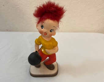 May include: A ceramic figurine of a boy with red hair, wearing a yellow shirt and red pants, holding a black bowling ball.