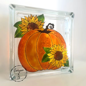 May include: A glass block with a painted design of a pumpkin and sunflowers. The pumpkin is orange and the sunflowers are yellow with brown centers. The glass block is clear and has a square shape.