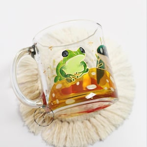 May include: A clear glass mug with a handle, featuring a hand-painted design of a green frog sitting on a mushroom. The mug is filled with a golden liquid and rests on a cream-colored coaster. The mug has a whimsical, artistic style.