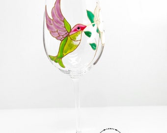 May include: A hand-painted wine glass with a hummingbird design. The hummingbird is green, pink, and gold, and is flying towards a white flower. The glass is clear and has a long stem.