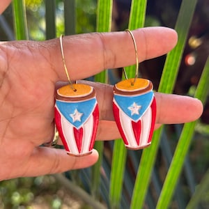 May include: A pair of gold hoop earrings with drum-shaped pendants. The pendants are painted with the Puerto Rican flag design: a blue triangle with a white star, red and white stripes, and a yellow top.