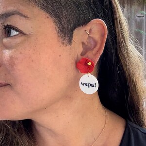 May include: A pair of earrings featuring a red flower charm and a white circle with the word "wepa!" in black lettering.