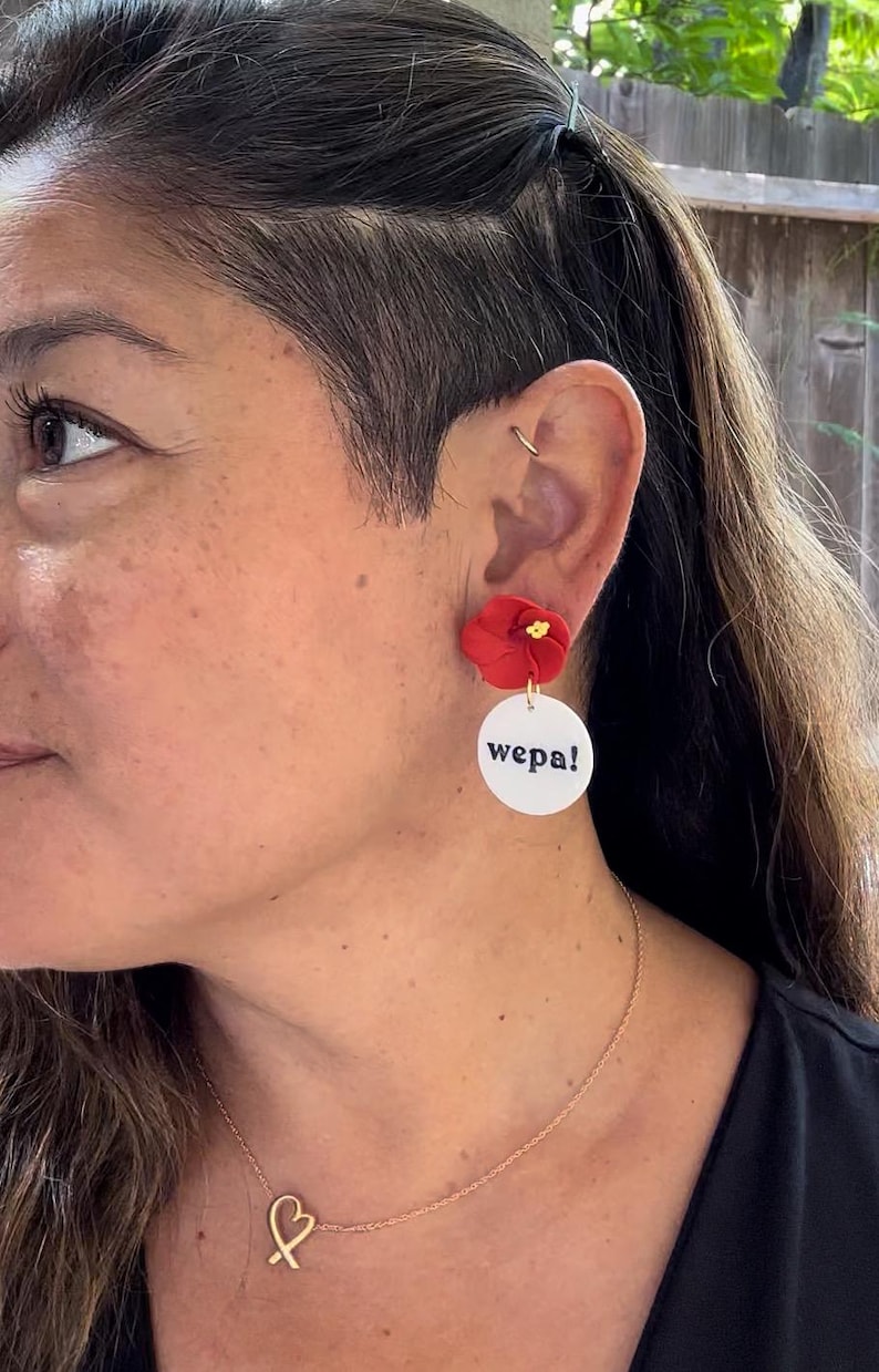 May include: A pair of earrings featuring a red flower charm and a white circle with the word "wepa!" in black lettering.