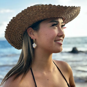 May include: A woman wearing a wide-brimmed straw hat and a black bikini top, looking off to the side. She is wearing gold hoop earrings with a pearl-like shell hanging from each earring.
