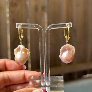 May include: A pair of gold hoop earrings with a large, pink baroque pearl dangling from each hoop.