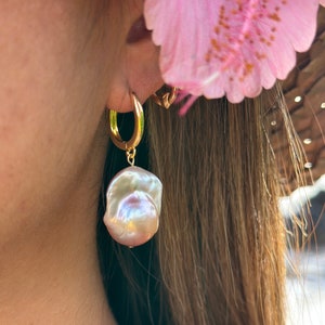 May include: Gold hoop earrings with a large, iridescent pearl drop. The pearl is a soft, off-white color with hints of pink and purple.
