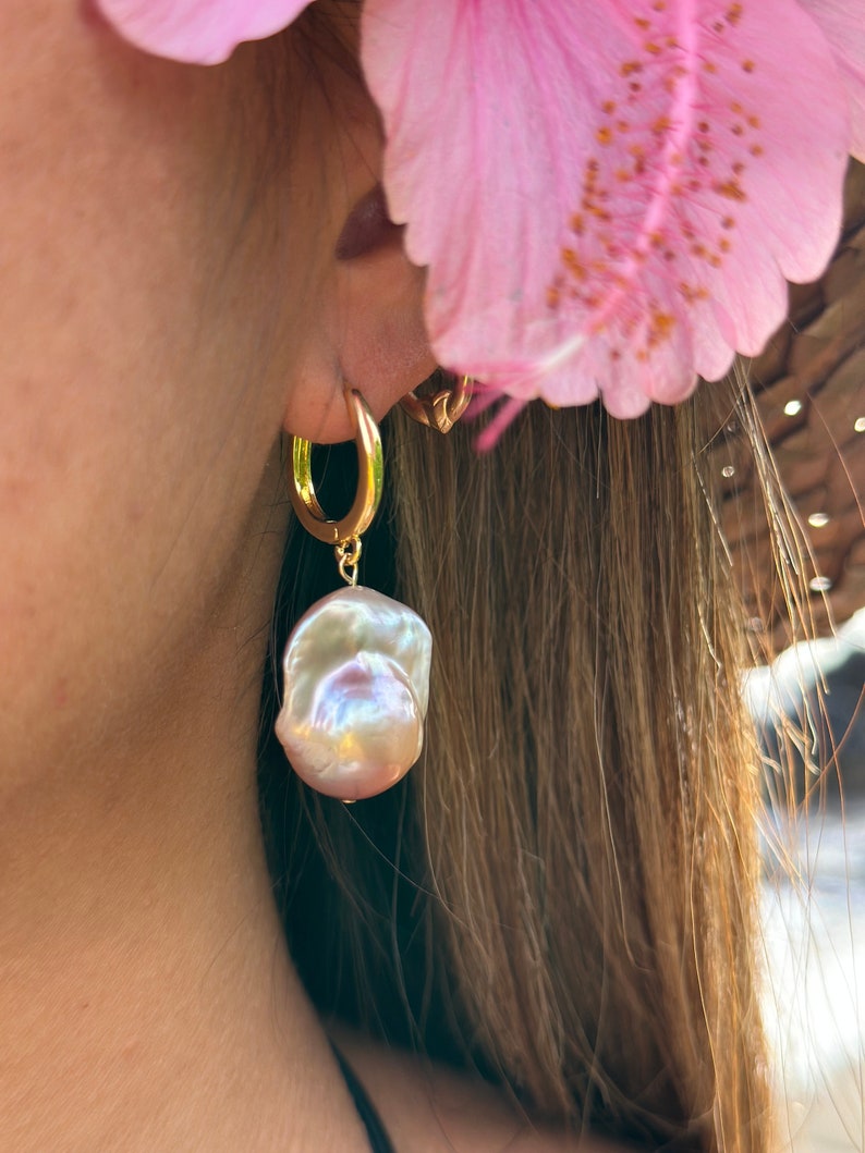 May include: Gold hoop earrings with a large, iridescent pearl drop. The pearl is a soft, off-white color with hints of pink and purple.