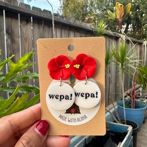 May include: A pair of red flower dangle earrings with white round charms that say "wepa!" in black text. The earrings are on a brown card that says "Made with Aloha".