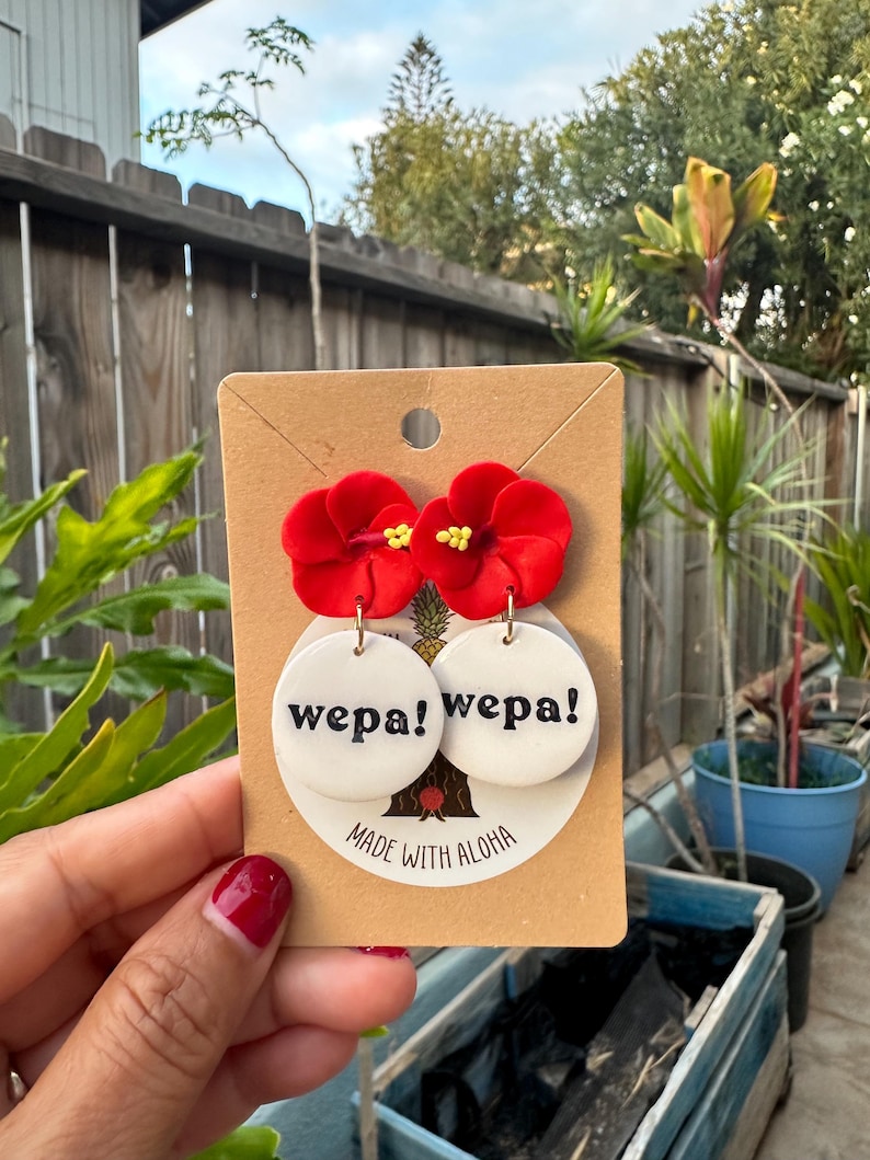 May include: A pair of red flower dangle earrings with white round charms that say "wepa!" in black text. The earrings are on a brown card that says "Made with Aloha".