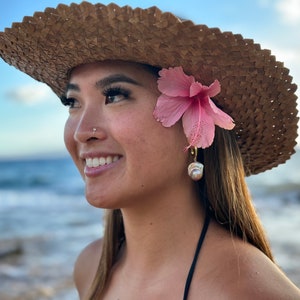 May include: A woman wearing a straw hat with a pink flower tucked behind her ear. She is wearing a pearl earring and has a nose ring. She is smiling and looking off to the side.