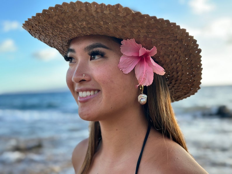 May include: A woman wearing a straw hat with a pink flower tucked behind her ear. She is wearing a pearl earring and has a nose ring. She is smiling and looking off to the side.