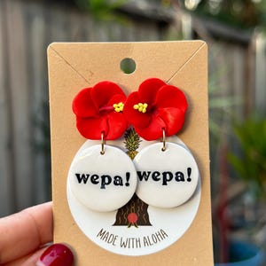 May include: Red flower dangle earrings with white round ceramic charms that say "wepa!" in black lettering. The earrings are on a brown card with the text "Made with Aloha".