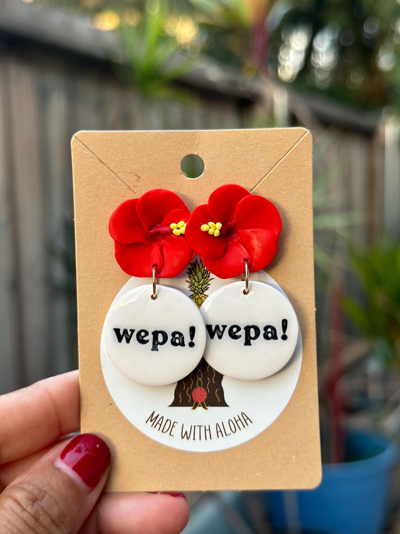 May include: Red flower dangle earrings with white round ceramic charms that say "wepa!" in black lettering. The earrings are on a brown card with the text "Made with Aloha".