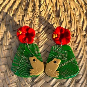 May include: A pair of green and gold frog earrings with red flower accents. The earrings are made of polymer clay and feature a detailed frog design with gold accents.