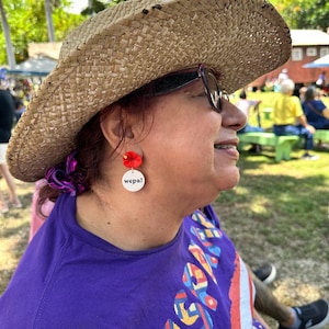 May include: A person wearing a straw hat and sunglasses with red flower earrings that say "wepa!" on the back. The person is wearing a purple shirt with a colorful design.