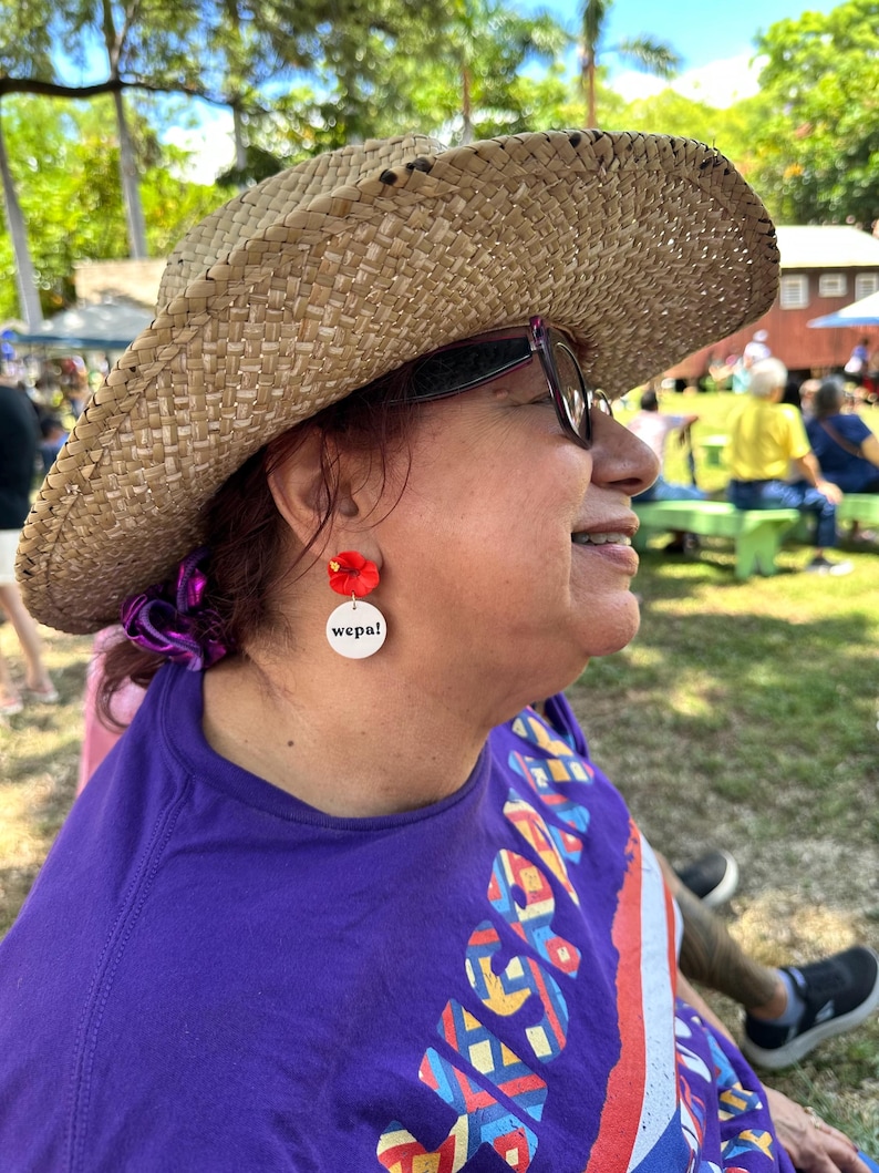 May include: A person wearing a straw hat and sunglasses with red flower earrings that say "wepa!" on the back. The person is wearing a purple shirt with a colorful design.