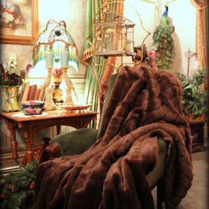 Op de afbeelding: Een bruine nep bont deken gedrapeerd over een pluche fauteuil in een vintage woonkamer. De deken is zacht en luxueus, met een subtiele glans. De tekst "Fur Accents Plush Pelt Throws" wordt weergegeven onder de deken.