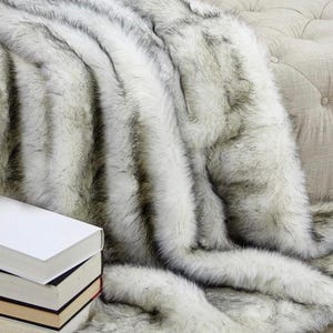 Pu&ograve; includere: Una coperta in pelliccia sintetica bianca e nera con un motivo a righe. La coperta &egrave; drappeggiata su un divano.
