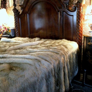 May include: A luxurious faux fur throw blanket in a light brown and gray color with a long pile. The blanket is draped over a wooden bed frame with a carved headboard.
