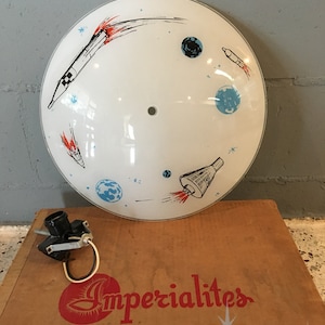 Op de afbeelding: Een witte, ronde lampenkap in mid-century modern stijl met een ruimtethema. Het heeft illustraties van raketten, planeten en sterren in zwart, rood en blauw. De kap staat bovenop een kartonnen doos met de tekst "Imperialites" en "Mid Century Metropolis".