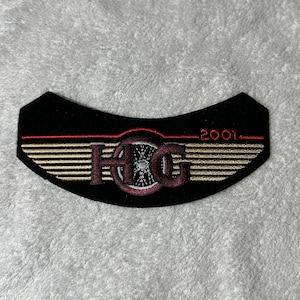 May include: A black, curved patch with gold and red embroidery. The design includes the letters "HOG" in a stylized font, a wheel design, and the year "2001". Gold wings are embroidered on either side of the central design.