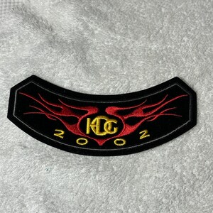 May include: Black, curved patch with red flame design and gold lettering. The patch features the letters "KDG" in gold, with the year "2002" below. The patch is likely for a motorcycle or biker-themed item.