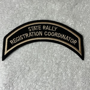 May include: A black and gold embroidered patch with the text "STATE RALLY REGISTRATION COORDINATOR" in gold lettering. The patch has a curved shape, possibly for a uniform or clothing item.