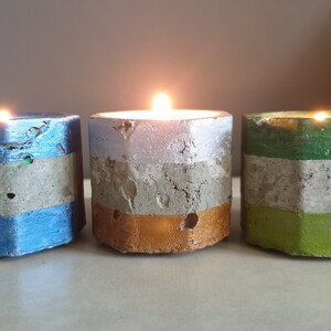 May include: Three lit tealight candles in hexagonal concrete holders. Each holder is painted with horizontal bands of colour: blue, silver, and green. The candles are placed on a light-coloured surface.