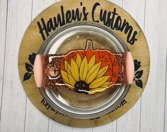 May include: A clear tumbler featuring a pumpkin and sunflower design. The pumpkin is orange with brown detailing, and the sunflower is yellow and brown. The tumbler has a glittery brown band and pink handles. It rests on a wooden coaster.