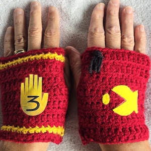 Pode incluir: Um par de luvas sem dedos de crochê vermelho com detalhes em amarelo e dourado. Uma luva tem uma mão amarela com o número "3" nela. A outra luva tem um design de Pac-Man amarelo.
