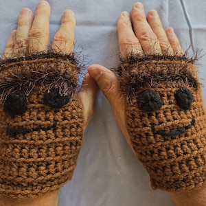 May include: A pair of brown crocheted fingerless gloves with a fuzzy brown trim and black embroidered eyes and a smiling mouth.