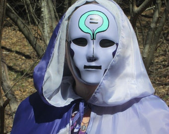 Peut inclure: Une personne portant une cape à capuche violette et blanche avec un masque blanc. Le masque a un symbole vert dessus.