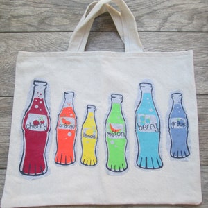 May include: A white canvas tote bag with a colorful design of six soda bottles. Each bottle has a different fruit flavor: cherry, orange, lemon, melon, berry, and grape.