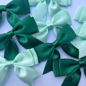 May include: A collection of satin bows in shades of green and mint green. The bows are displayed on a white surface, with different shades of green creating a contrast. These bows are suitable for various craft projects.
