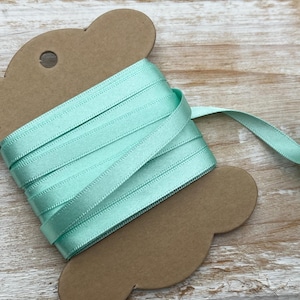 May include: A spool of light green satin ribbon on a brown cardstock tag. The ribbon is wound around the tag and has a slight sheen.
