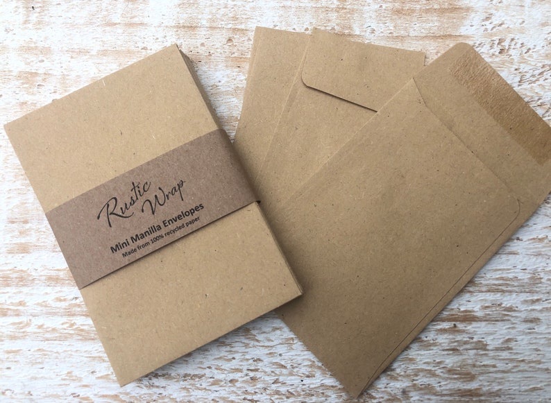 Op de afbeelding: Een stapel bruine mini manila enveloppen gemaakt van 100% gerecycled papier. De enveloppen zijn verpakt met een bruin bandje waarop "Rustic Wrap" en "Mini Manila Envelopes" staat.