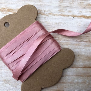 May include: A spool of pale pink satin ribbon wound around a brown cardboard spool. The ribbon is smooth and shiny.