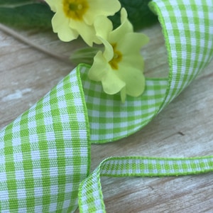 May include: A close-up of a green and white gingham ribbon, with a delicate yellow flower and green leaves in the background. The ribbon is curled and twisted, showing its pattern. The flowers have yellow petals and a yellow centre.