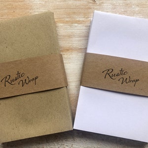 Op de afbeelding: Twee stapels bruin cadeaupapier met een bruin lint om elke stapel. Het lint heeft de tekst "Rustic Wrap" erop gedrukt.