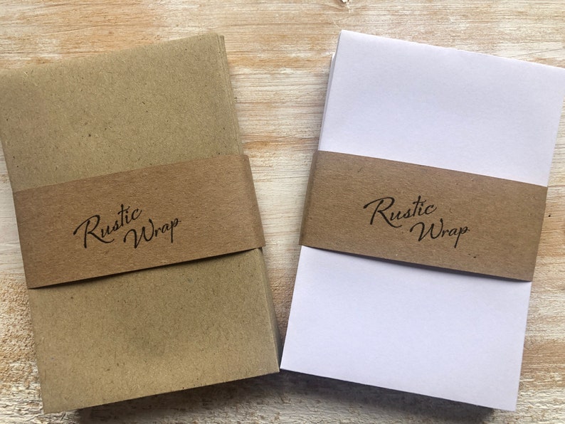 Op de afbeelding: Twee stapels bruin cadeaupapier met een bruin lint om elke stapel. Het lint heeft de tekst "Rustic Wrap" erop gedrukt.